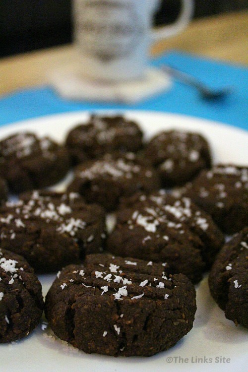 Several chocolate cookies sprinkled with coconut and placed on a white plate. A coffee mug and teaspoon can be seen in the background.