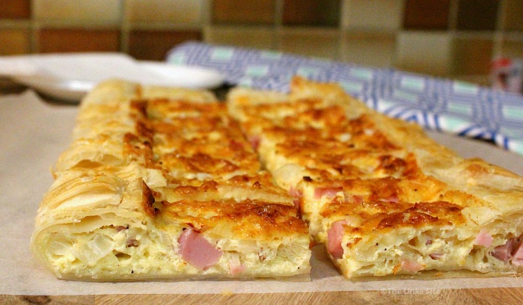 Wide angle picture showing slices of quiche on a wooden board with a green and blue patterned tea towel in the background.