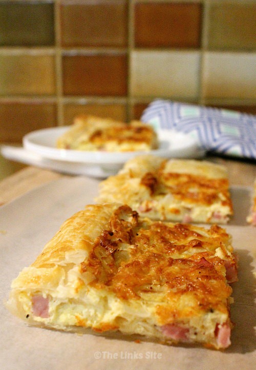 Two pieces of puff pastry quiche can be seen on a wooden board. Another slice of quiche on a white plate and be seen in the background along with a green and blue patterned tea towel.