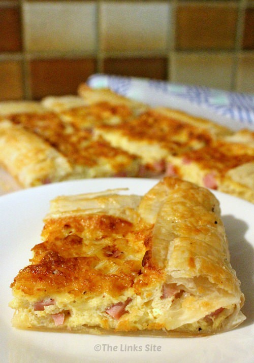 Slice of puff pastry quiche on a white plate. More slices of quiche and a tea towel can be seen in the background.