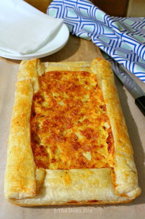 Overhead view of rectangular quiche before slicing. The quiche is on a wooden board and there are white plates and a green and blue patterned tea towel in the background. 