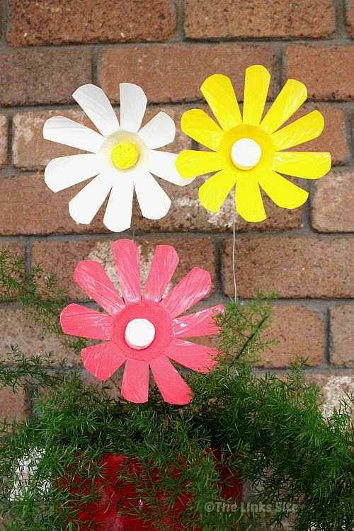 A pink, a white, and a yellow plastic bottle flower sticking up out of a potted plant. There is a brick wall in the background.
