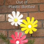 A pink, a white, and a yellow plastic bottle flower sticking up out of a potted plant. There is a brick wall in the background. Text overlay says: How to Make Flowers Out of Plastic Bottles!