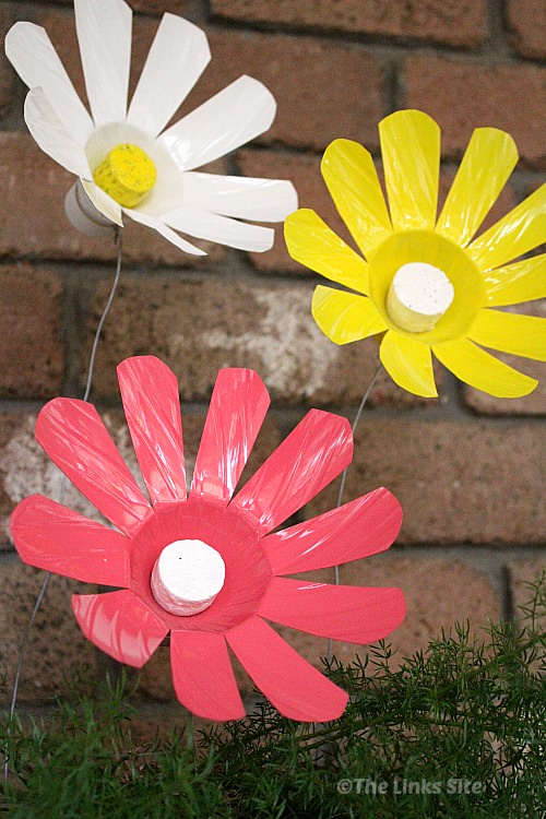A pink, a white, and a yellow plastic bottle flower sticking up out of a plant pot on wires. A brick wall can be seen in the background.