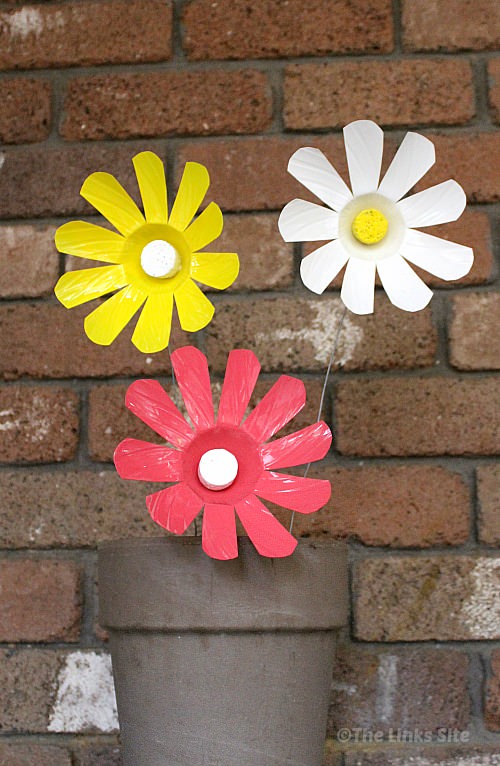 A pink, a white, and a yellow plastic bottle flower sticking up our of an empty grey clay pot with a brick wall in the background.