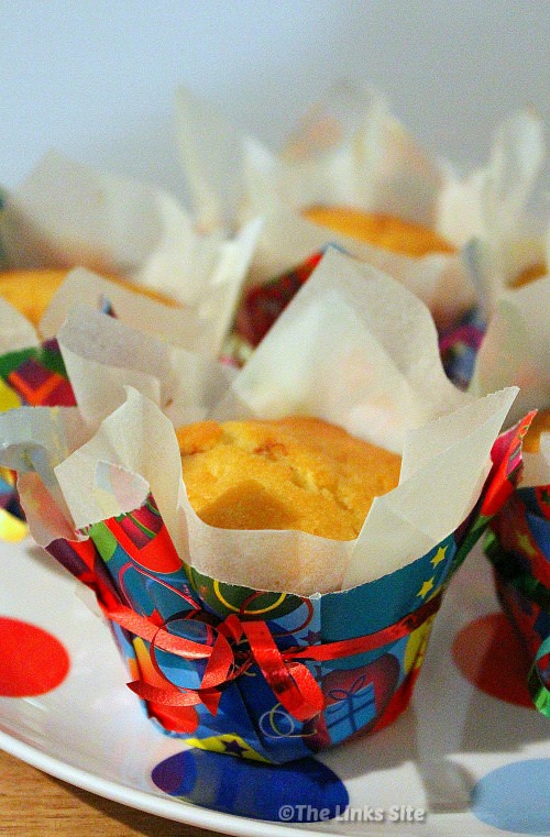 Several muffins on a white plate with multi coloured spots. Each muffin is in a baking paper liner and then an outer liner made from multi coloured wrapping paper. Curling ribbon is tied around the muffins for decoration.