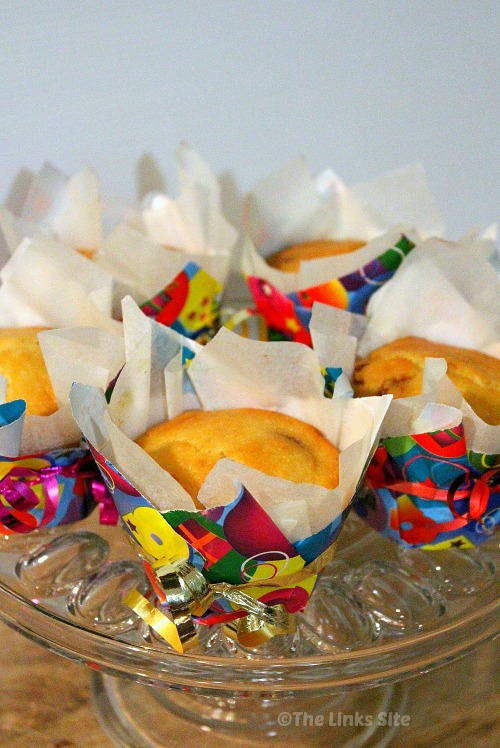 A group of chocolate chip muffins on a glass cake stand. Each muffin is in a baking paper liner and then an outer liner made from multi coloured wrapping paper. Curling ribbon is tied around the muffins for decoration.