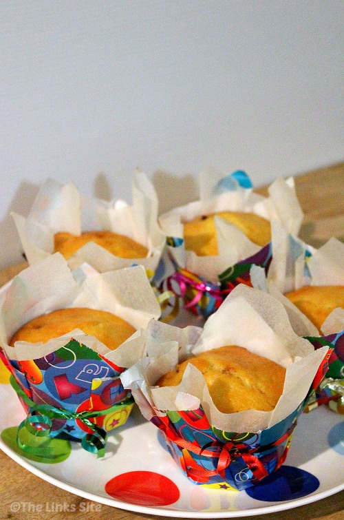 Five chocolate chip muffins on a white plate with multi coloured spots. Each muffin is in a baking paper liner and then an outer liner made from multi coloured wrapping paper. Curling ribbon is tied around the muffins for decoration.