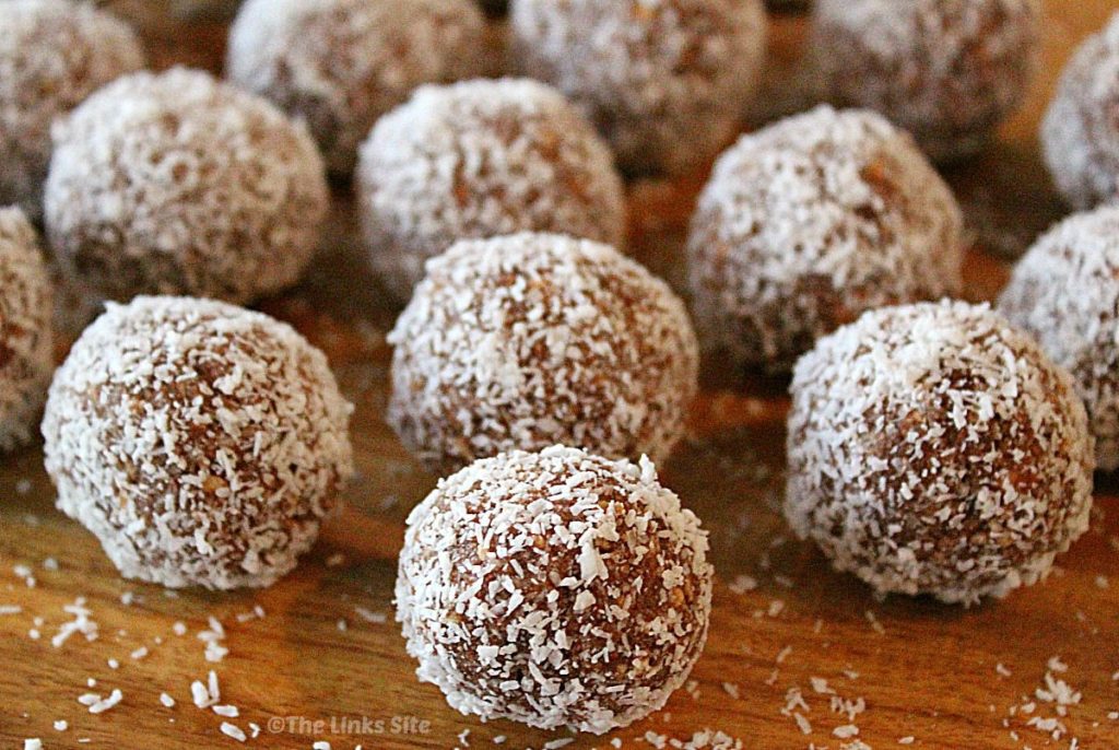 Lots of coconut coated balls are arranged on a wooden board with small specks of coconut scattered around the balls. One central ball is in focus in the foreground and several other balls can be seen to the side and in the background.