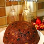 Whole traditional Christmas plum pudding on a white plate. A wine bottle, glasses, and Christmas decorations can be seen in the background. Text overlay says: Easy Steamed Christmas Pudding!