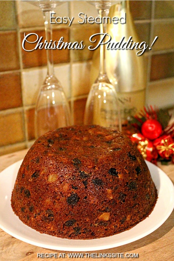 Whole traditional Christmas plum pudding on a white plate. A wine bottle, glasses, and Christmas decorations can be seen in the background. Text overlay says: Easy Steamed Christmas Pudding!