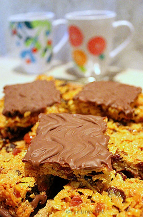 A pile of Florentine squares on a wooden board where the top few squares are sitting chocolate side up. There are two mugs in the background