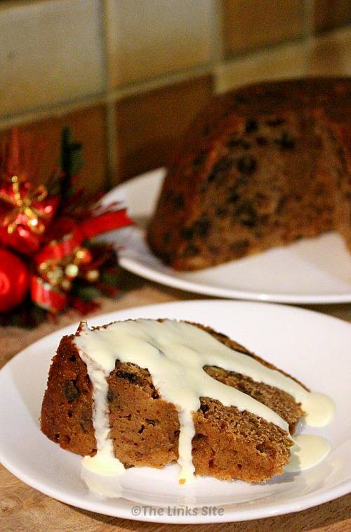 Slice of plum pudding topped with cream on a white plate. The rest of the pudding and some festive decorations can be seen in the background.