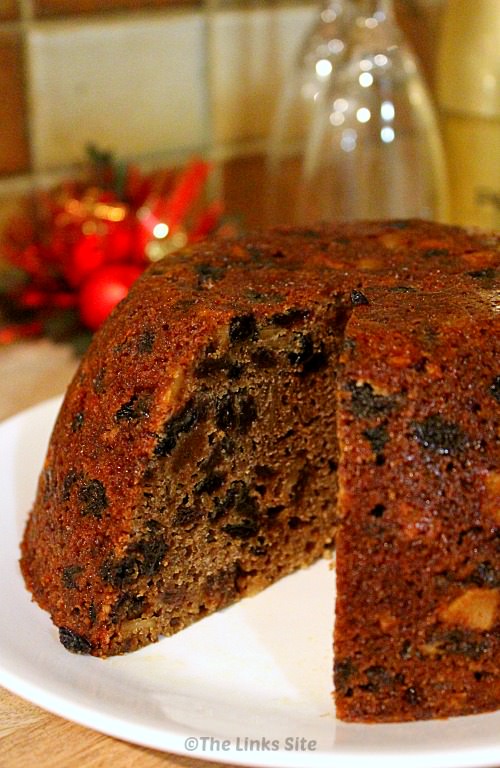 Traditional plum pudding on a white plate with a piece removed. Festive decorations can be seen in the background.