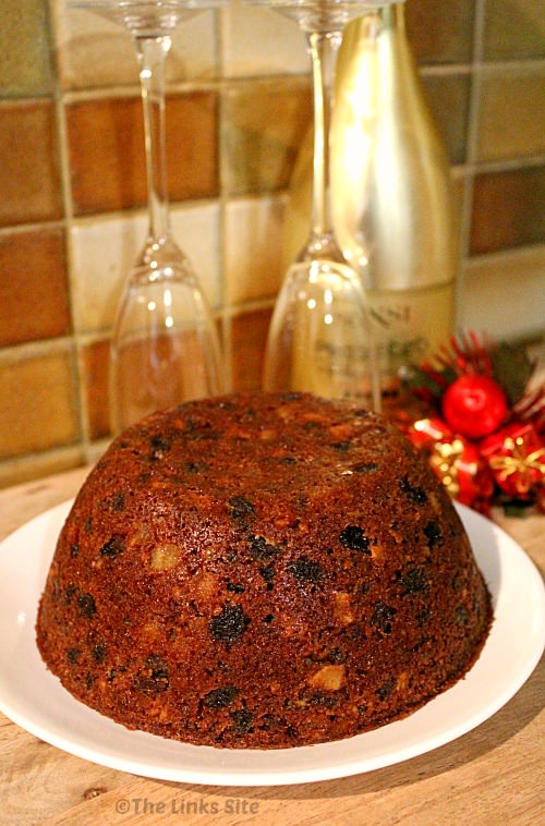Whole traditional Christmas plum pudding on a white plate. A wine bottle, glasses, and Christmas decorations can be seen in the background.