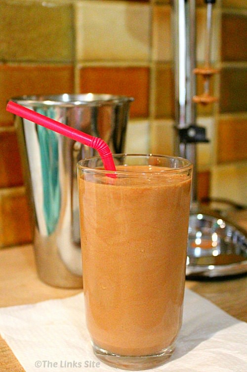 Snickers milkshake in a tall glass with a straw. A metal milkshake cup and milkshake maker can be seen in the background.