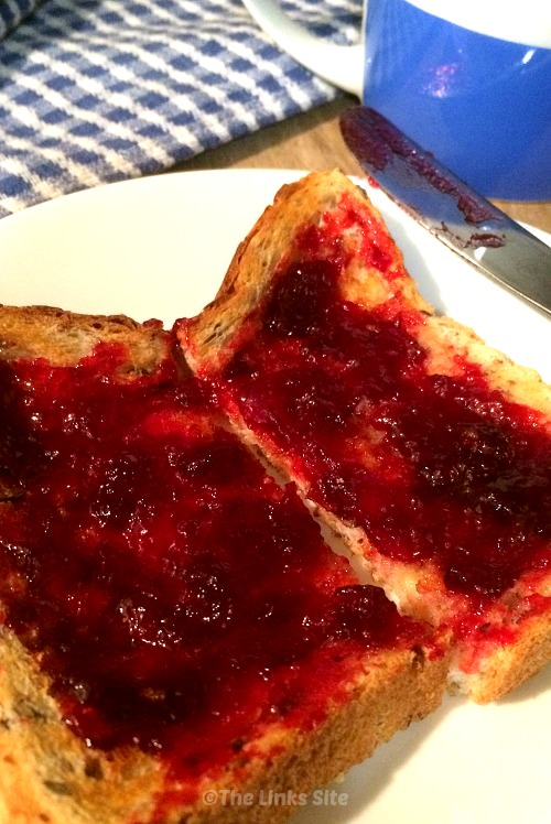 Slice of multigrain toast with plum jam on white plate with knife, mug, and tea towel in the background.