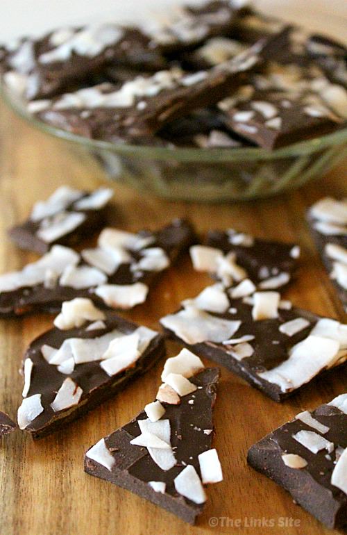 Broken up pieces of chocolate bark topped with coconut flakes on a wooden board. In the background there is a glass bowl filled with more chocolate bark.