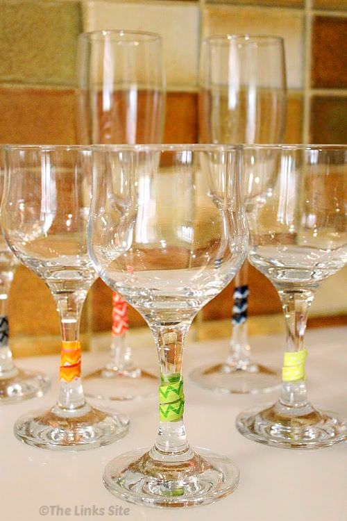 Several stemmed wine glasses are arranged on a white mat. The stems are decorated with colourful wraps.