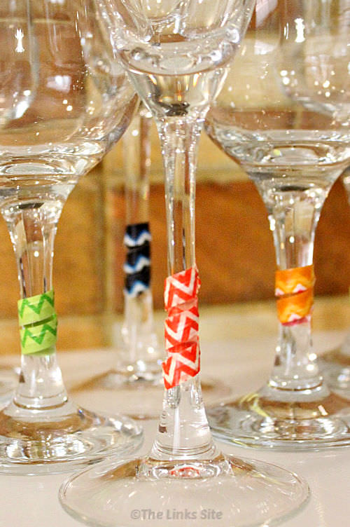 Close up of the stem of four wine glasses. Brightly coloured washi tape covered twist ties have been wrapped around each wine glass stem.