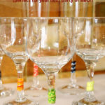 Several stemmed wine glasses are arranged on a white mat. The stems are decorated with colourful wraps. Text overlay says: Easy DIY Wine Stem Wraps (perfect for your next party or BBQ!).
