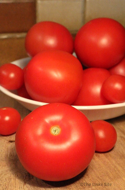 A bowl of ripe red tomatoes of various sizes on a wooden cutting board. A couple of tomatoes are placed on the board in front of the bowl.