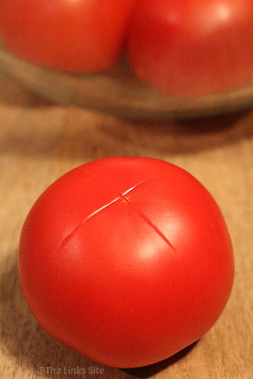 A single ripe tomato with a shallow X cut into the bottom is placed on a wooden cutting board. A glass bowl containing more tomatoes can be seen in the background.