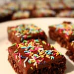 Several pieces of chocolate slice are placed on a white plate, with only the front piece fully in focus. More pieces of slice can be seen further in the background. The pieces of slice are topped with colourful sprinkles.