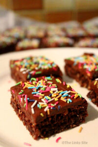 Several pieces of chocolate slice are placed on a white plate, with only the front piece fully in focus. More pieces of slice can be seen further in the background. The pieces of slice are topped with colourful sprinkles.
