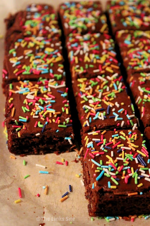 The chocolate coconut slice is pictures on the baking paper it was cooked on. It has been topped with chocolate icing and lots of colourful sprinkles and cut into squares. Extra sprinkles are scattered around on the baking paper.