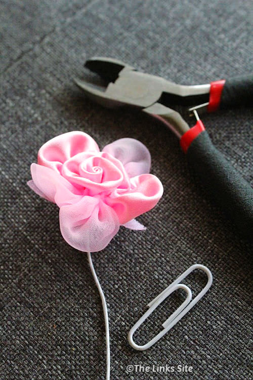A pink craft flower, a white paperclip, and a pair of plyers resting on grey fabric.