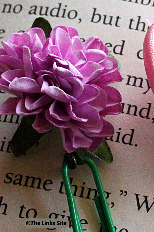 Close up of a green bookmark with a purple flower on the page of a book.
