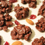 Several chocolate clusters are arranged on a piece of baking papers with dried cranberries and almonds scattered around them.