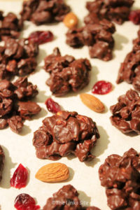 Several chocolate clusters are arranged on a piece of baking papers with dried cranberries and almonds scattered around them.