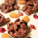 A single chocolate cluster is seen in the foreground with cranberries and almonds scattered around it. More clusters can be seen in the background.
