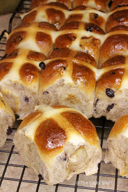 Hot cross buns cooling on a rack. Several buns have been separated while the rest of the still joined buns can be seen in the background.