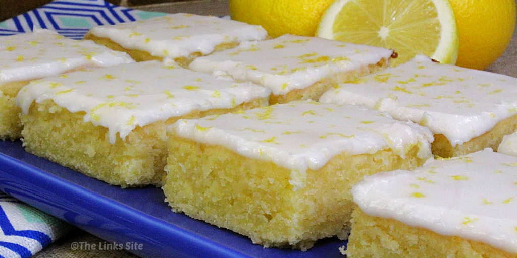 Iced lemon brownies, arranged in two rows on a blue plate. A blue and white patterned tea towel, half a lemon, and two whole lemons can be seen in the background.