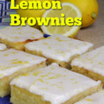 Iced lemon brownies on a blue plate. A blue and white patterned tea towel, two whole lemons, and a half of a lemon can be seen in the background. Text overlay says: Deliciously Tangy & Fudgy Lemon Brownies.