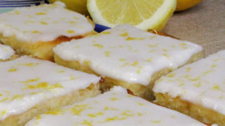 Iced lemon brownies on a blue plate. A blue and white patterned tea towel, two whole lemons, and a half of a lemon can be seen in the background.