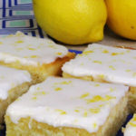 Iced lemon brownies on a blue plate. A blue and white patterned tea towel and two lemons can be seen in the background.