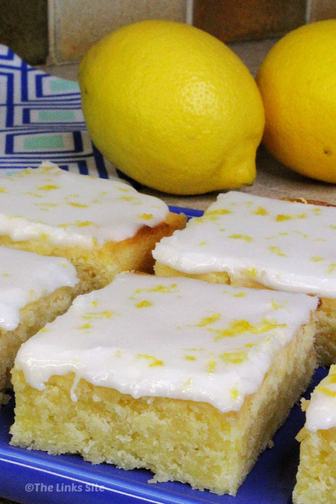 Iced lemon brownies on a blue plate. A blue and white patterned tea towel and two lemons can be seen in the background.