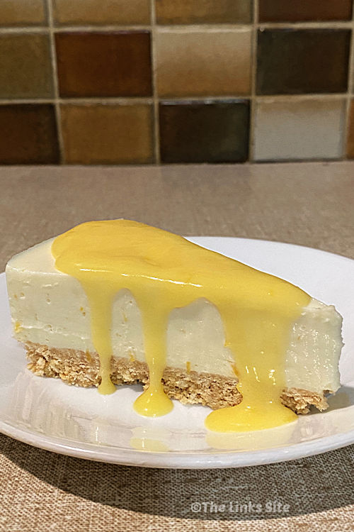A piece of cheesecake is pictured on a white plate. The cheesecake has been topped with some lemon curd which is dripping down the sides.