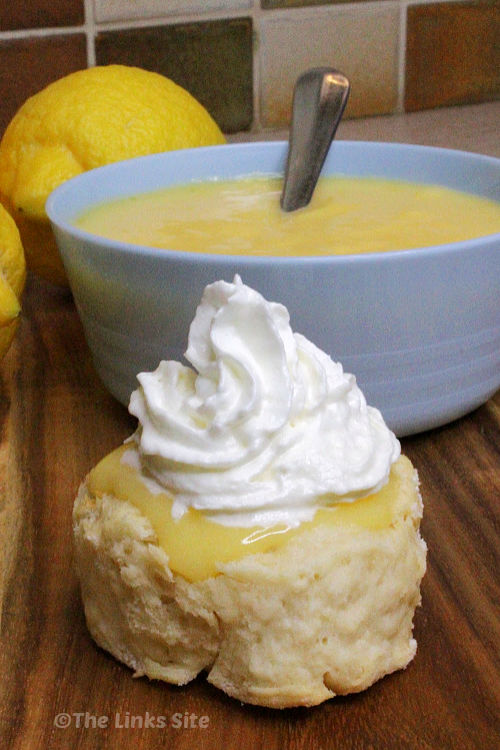 Half of a scone has been topped with lemon curd and then whipped cream. A blue bowl containing more lemon curd can be seen in the background along with some lemons.