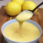 Lemon curd is dripping from a spoon into a Pale blue bowl containing more lemon curd. The bowl is on a wooden board and three lemons can be seen in the background.