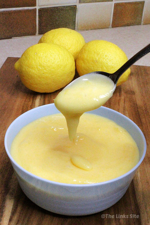 Lemon curd is dripping from a spoon into a Pale blue bowl containing more lemon curd. The bowl is on a wooden board and three lemons can be seen in the background.