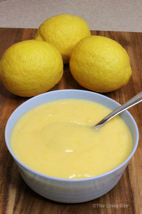 A spoon is pictured in a bowl of lemon curd. The bowl is on a wooden board and three lemons can be seen in the background.