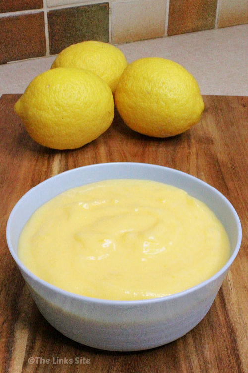 Pale blue bowl containing lemon curd pictured on a wooden board. Three lemons can be seen in the background.