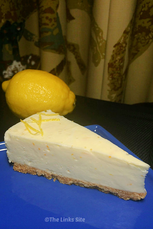 One wedge shaped slice of cheesecake on a blue plate with a whole lemon in the background.
