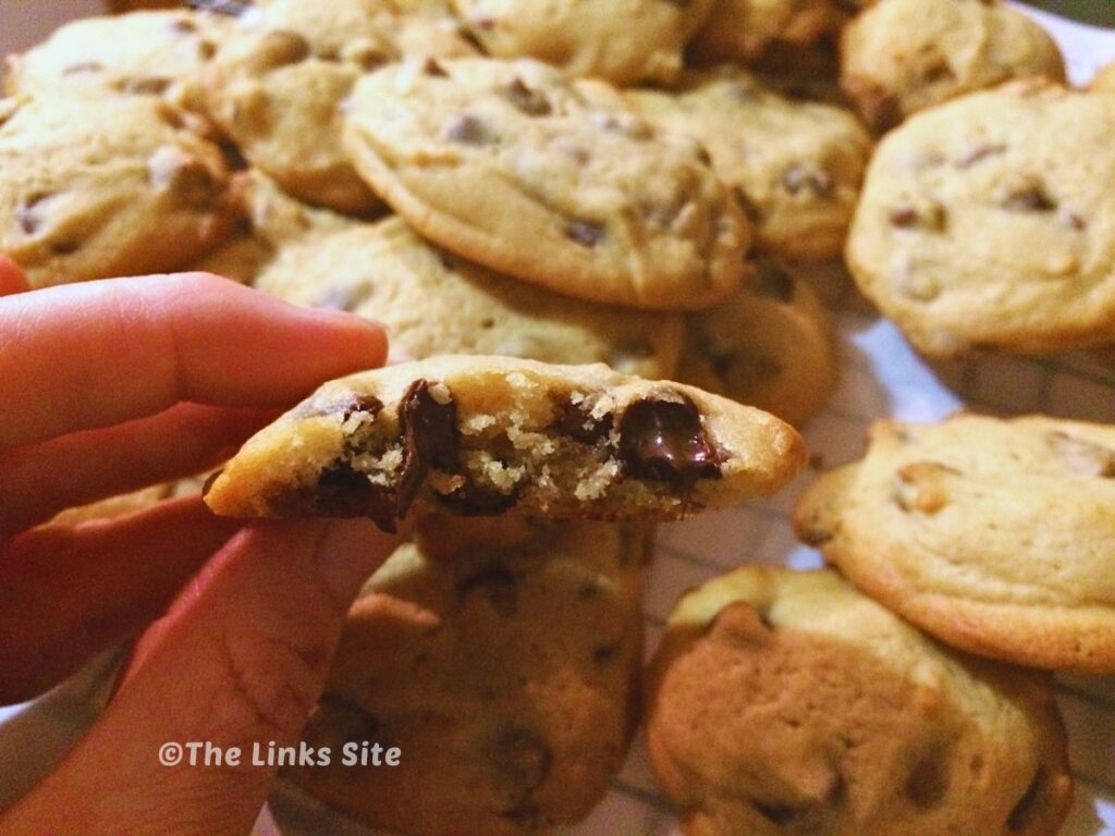 One, still warm, caramel chocolate chip cookie is broken in half to show soft gooey chocolate and caramel chips with a plate with more cookies can be seen in the background.