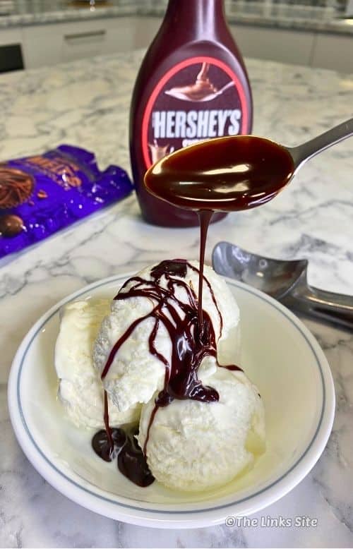 Spoon drizzling warm hot fudge sauce over vanilla ice cream in a white bowl, with Hershey’s syrup, chocolate buttons and an ice-cream scoop in the background.